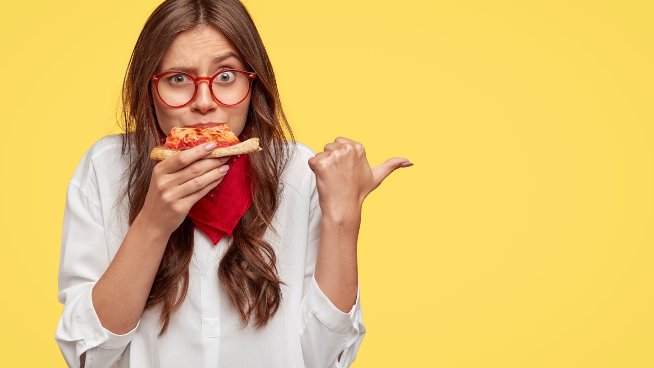 horizontal-shot-of-surprised-female-eats-tasty-slice-of-pizza-dressed-in-fashionable-clothes-indicates-with-thumb-invites-you-to-pizzeria-isolated-over-yellow-wall-people-and-nutrition