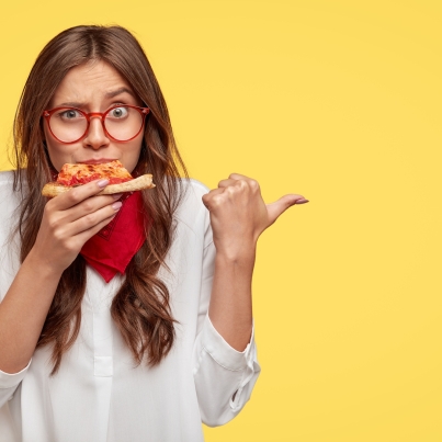 horizontal-shot-of-surprised-female-eats-tasty-slice-of-pizza-dressed-in-fashionable-clothes-indicates-with-thumb-invites-you-to-pizzeria-isolated-over-yellow-wall-people-and-nutrition
