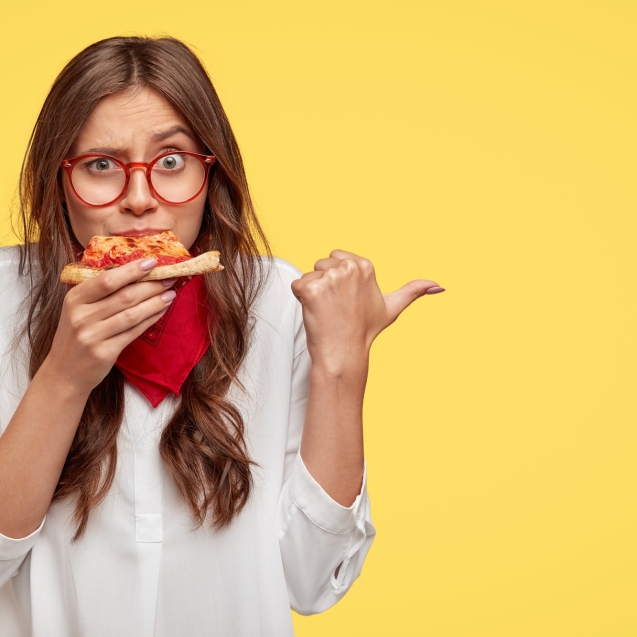 horizontal-shot-of-surprised-female-eats-tasty-slice-of-pizza-dressed-in-fashionable-clothes-indicates-with-thumb-invites-you-to-pizzeria-isolated-over-yellow-wall-people-and-nutrition