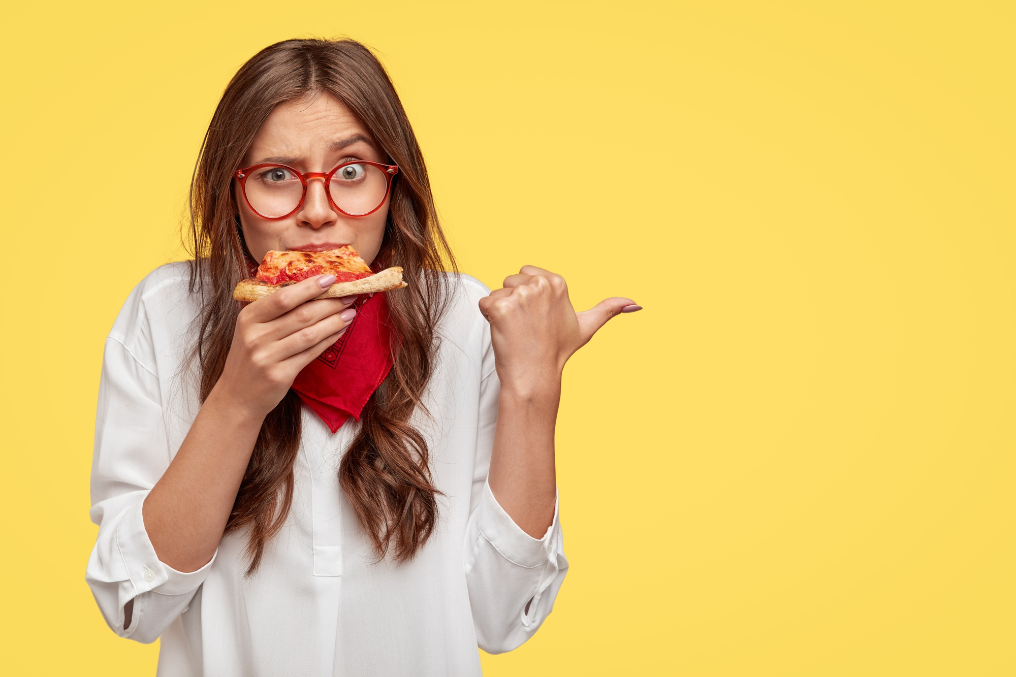 horizontal-shot-of-surprised-female-eats-tasty-slice-of-pizza-dressed-in-fashionable-clothes-indicates-with-thumb-invites-you-to-pizzeria-isolated-over-yellow-wall-people-and-nutrition