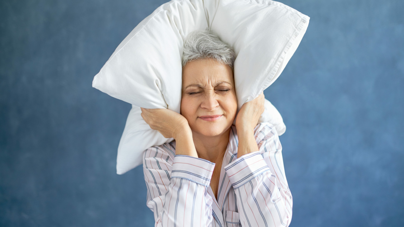 Annoyed dissatisfied mature woman with gray hair grimacing having sleepless night because of loud music, snoring husband, covering ears with pillow, having angry irritated facial expression