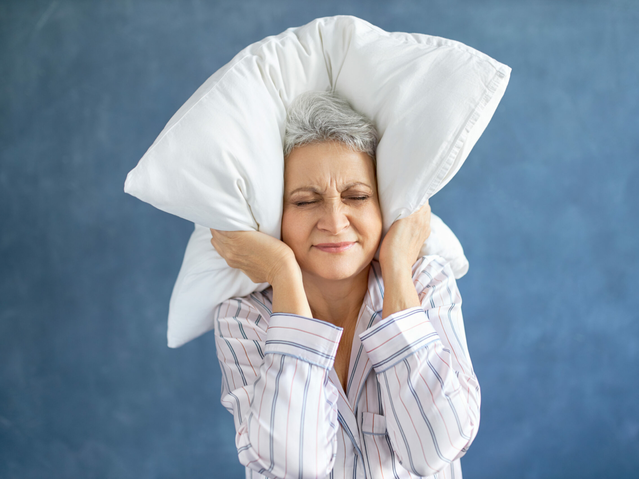 Annoyed dissatisfied mature woman with gray hair grimacing having sleepless night because of loud music, snoring husband, covering ears with pillow, having angry irritated facial expression