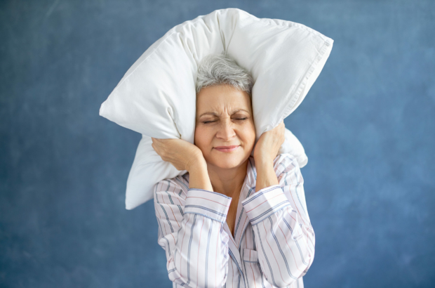 Annoyed dissatisfied mature woman with gray hair grimacing having sleepless night because of loud music, snoring husband, covering ears with pillow, having angry irritated facial expression